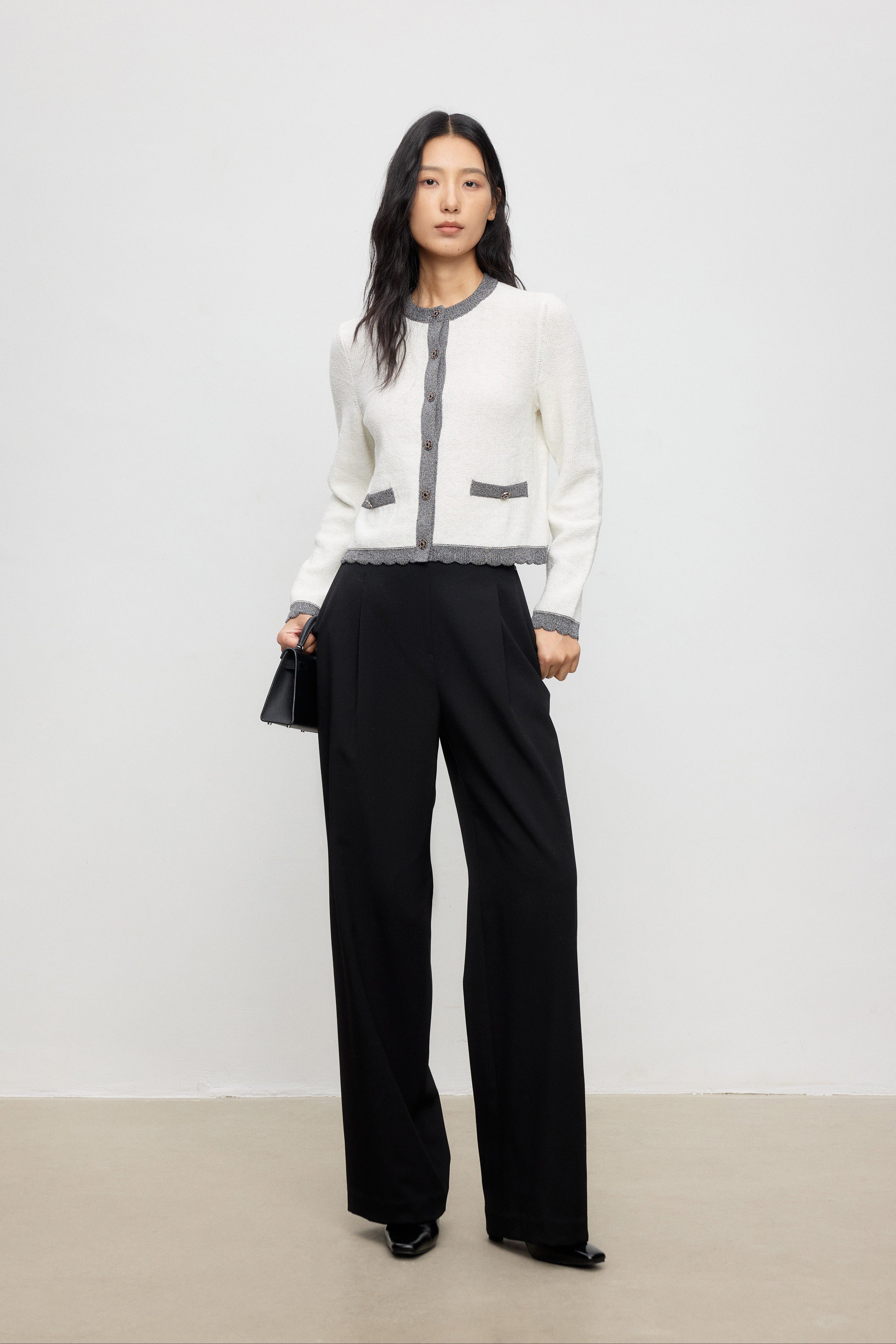 Woman wearing a white cardigan with gray accents and black pants on a plain background