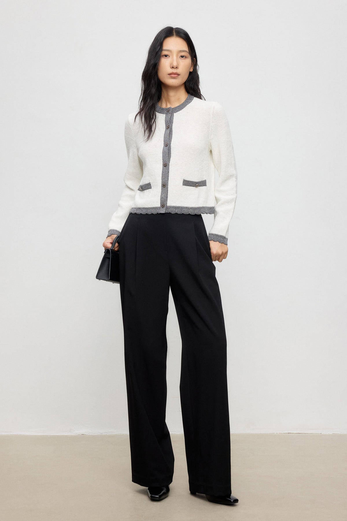 Woman wearing a white cardigan with gray accents and black pants on a plain background