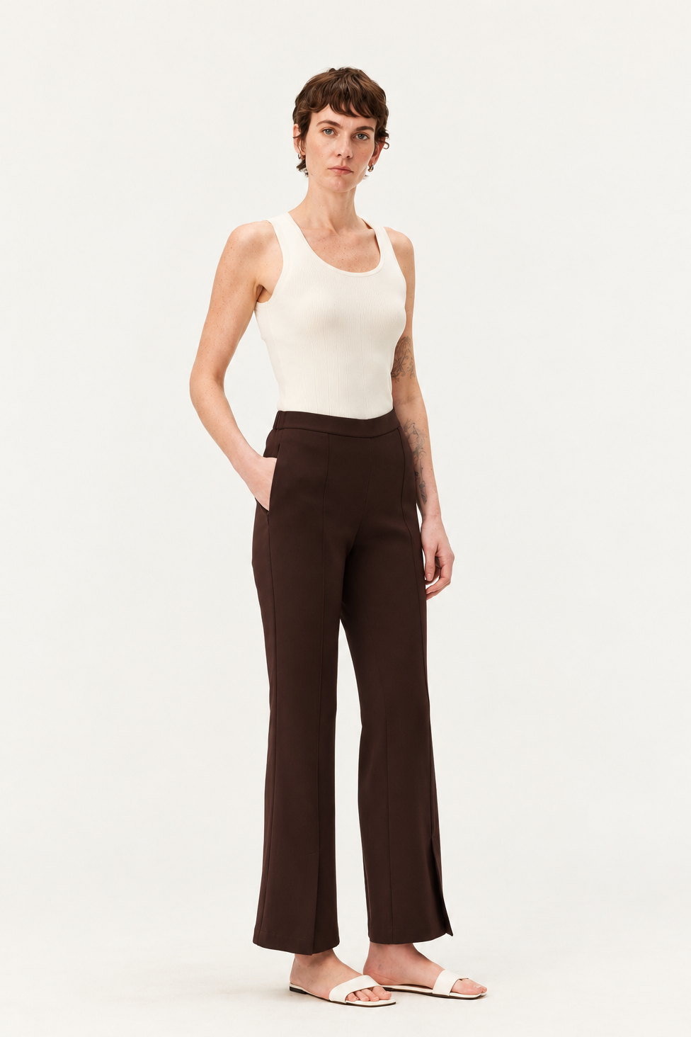 A model stands against a plain white studio background wearing dark brown wide-leg cropped pants, a cream ribbed tank top, and white slide sandals.