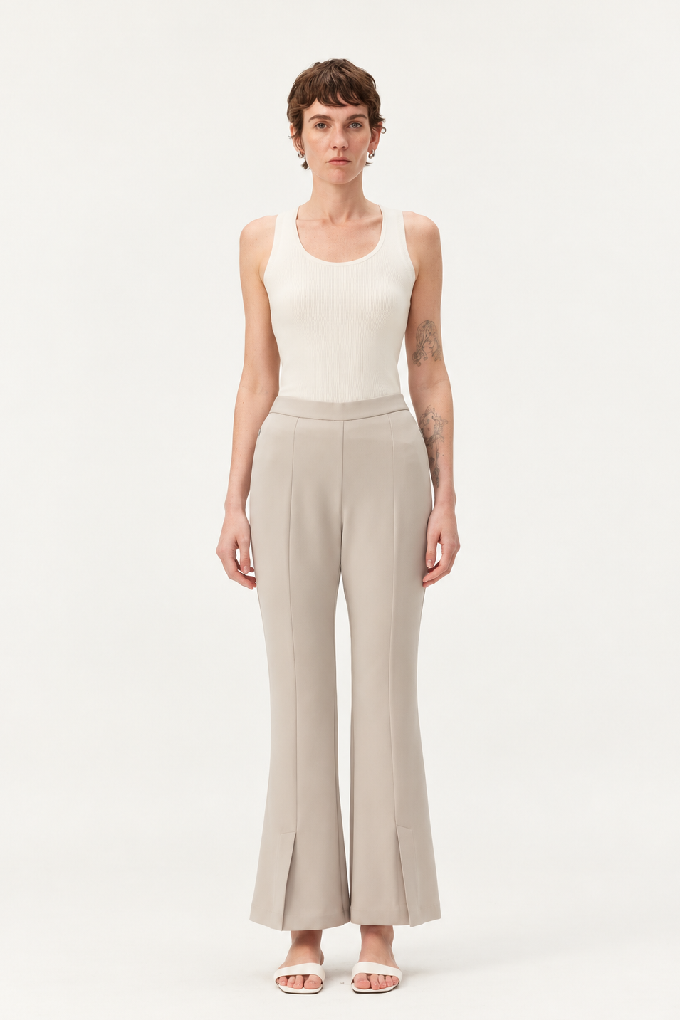 A model stands against a plain white studio background wearing a sleeveless cream top and light beige wide-leg trousers with a high waist and subtle front slit detail at the hem, paired with white flat sandals.