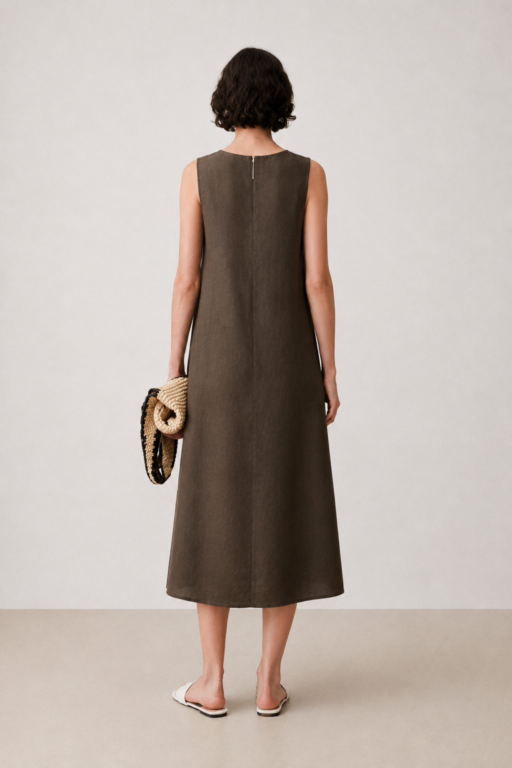 A woman stands with her back to the camera wearing a sleeveless, knee-length brown dress. She is holding a woven clutch and wearing white slide sandals, set against a neutral studio background.