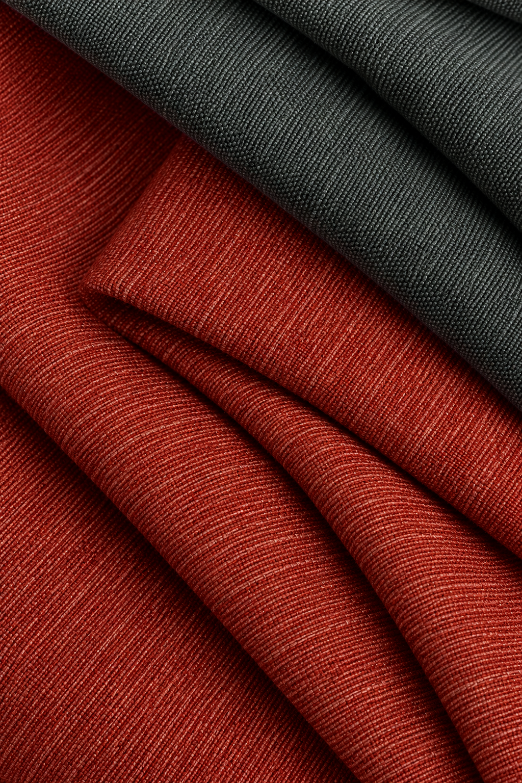 Folded textured fabric in two colors: a rust red fabric in the foreground and a dark charcoal gray fabric in the background, both showing a fine ribbed weave.