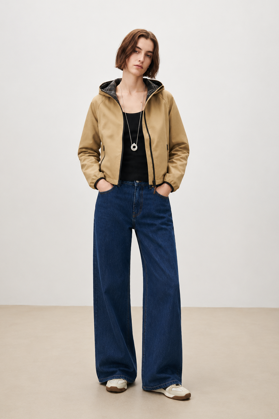 A model stands in a studio wearing a tan hooded jacket with a black interior lining, a black top, wide-leg dark blue jeans, and white sneakers.
