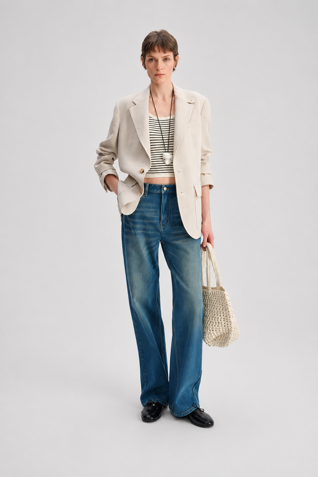 A model stands against a light gray studio background wearing a beige blazer over a black-and-white striped crop top, blue wide-leg jeans, black shoes, and holding a woven straw tote bag.