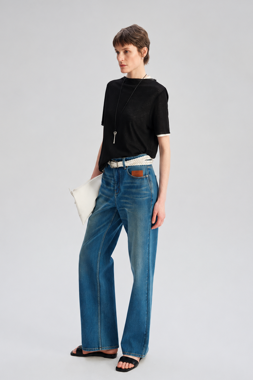 A model stands against a light gray studio background wearing blue wide-leg jeans, a black short-sleeve top, a white belt, black sandals, and holding a white clutch.