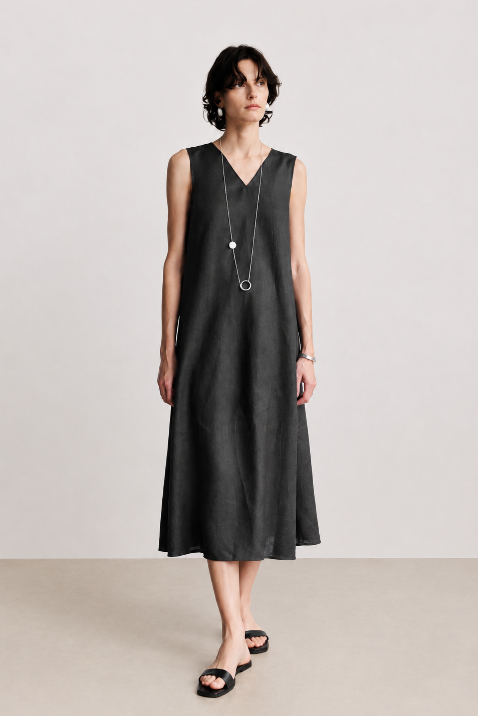 A woman modeling a sleeveless black midi dress with a V-neckline, styled with a long silver pendant necklace and black slide sandals against a neutral studio backdrop.