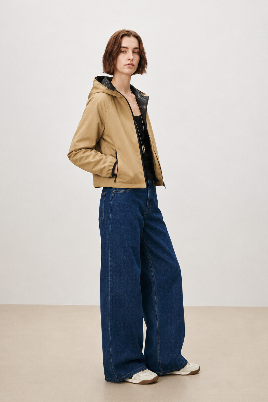 A model stands in a studio wearing a tan hooded jacket over a black top, paired with wide-leg dark blue jeans and white sneakers.