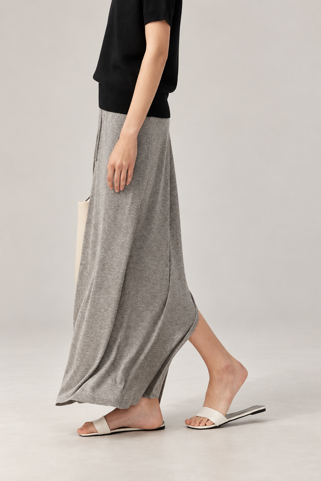 A model wearing a long, flowing gray maxi skirt paired with a black short-sleeve top and white slide sandals, standing against a light gray studio background.