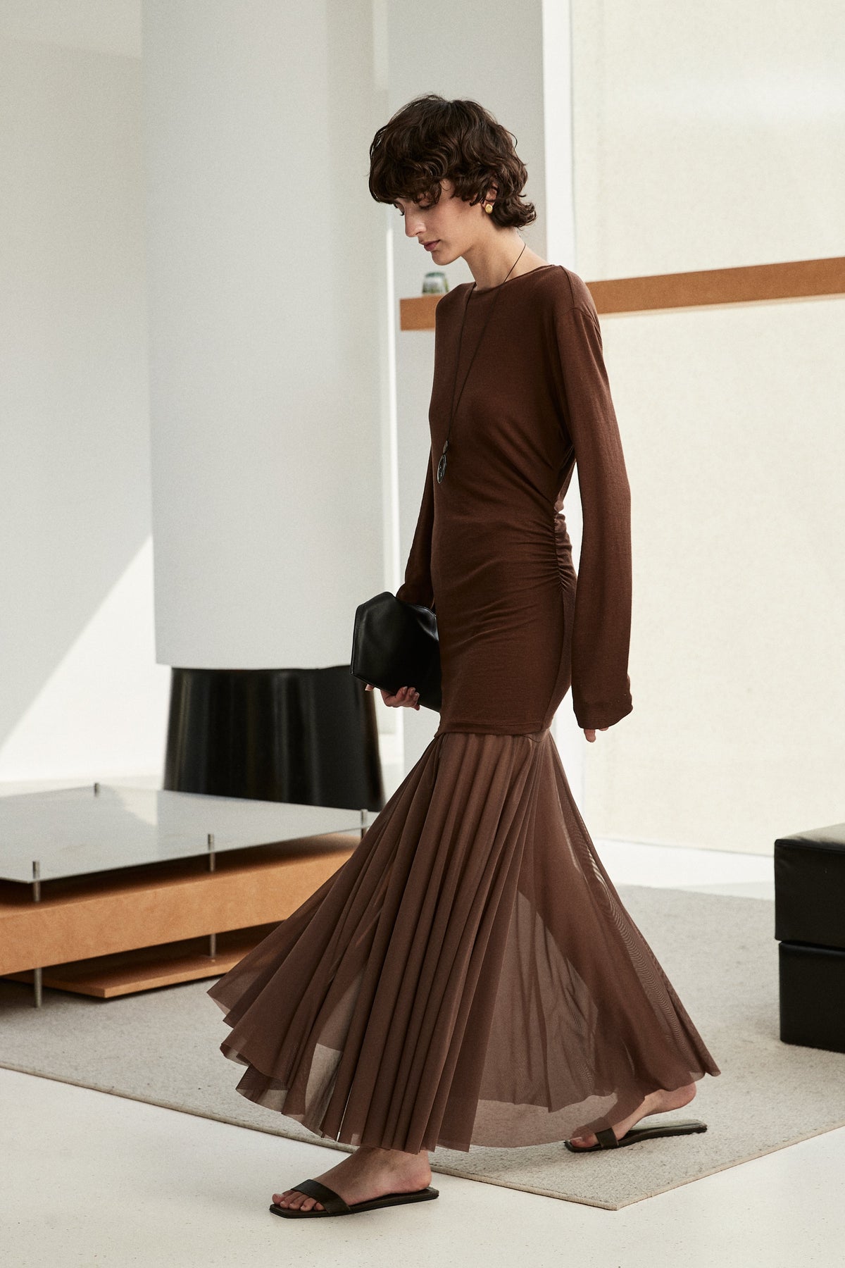 A woman wearing a long-sleeved brown ribbed knit top and a matching pleated midi skirt, walking in a minimalist modern interior with white walls and neutral furniture.