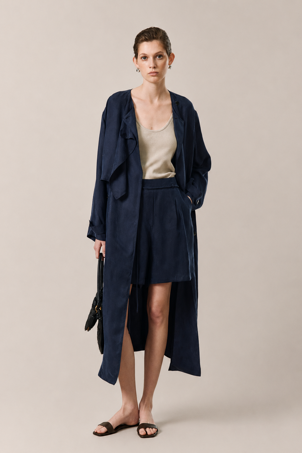A model stands against a plain beige studio background wearing a navy blue long drape coat over a beige top and matching navy skirt, holding a black handbag.