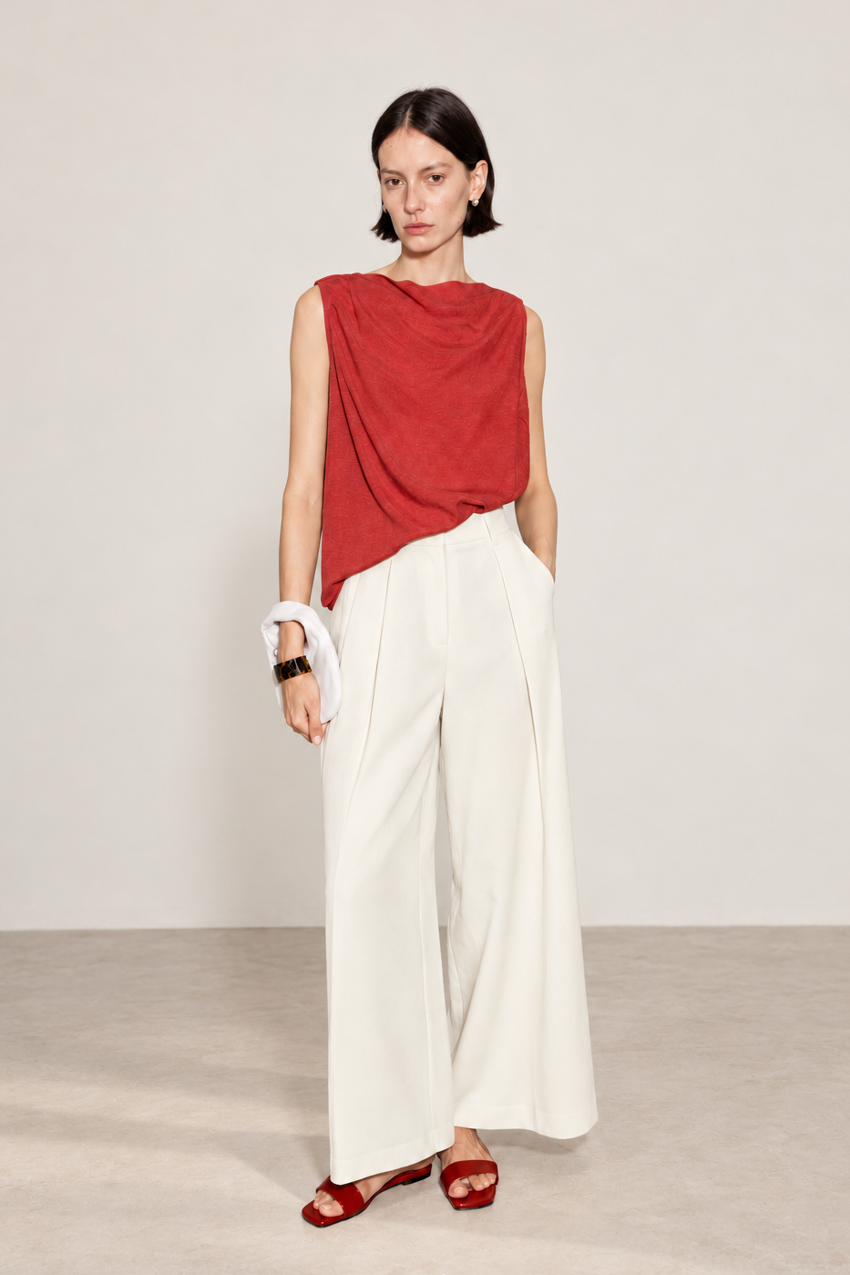 A model stands in a studio wearing a sleeveless draped red top, high-waisted white wide-leg trousers, and red sandals, with a white clutch in one hand.