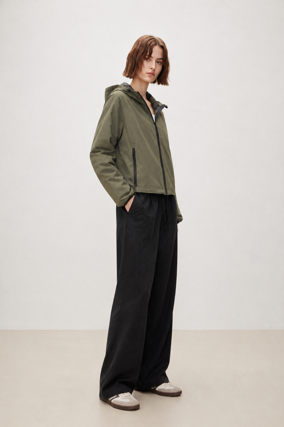 A model stands in a studio wearing an olive green hooded zip-up jacket, black wide-leg trousers, and white slip-on shoes against a plain light background.