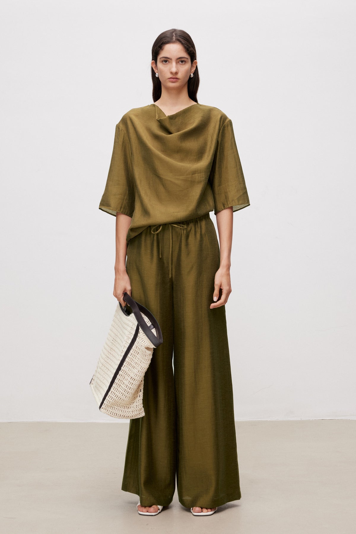 Woman wearing an olive green outfit with wide-leg pants and a top, holding a woven bag on a plain background.