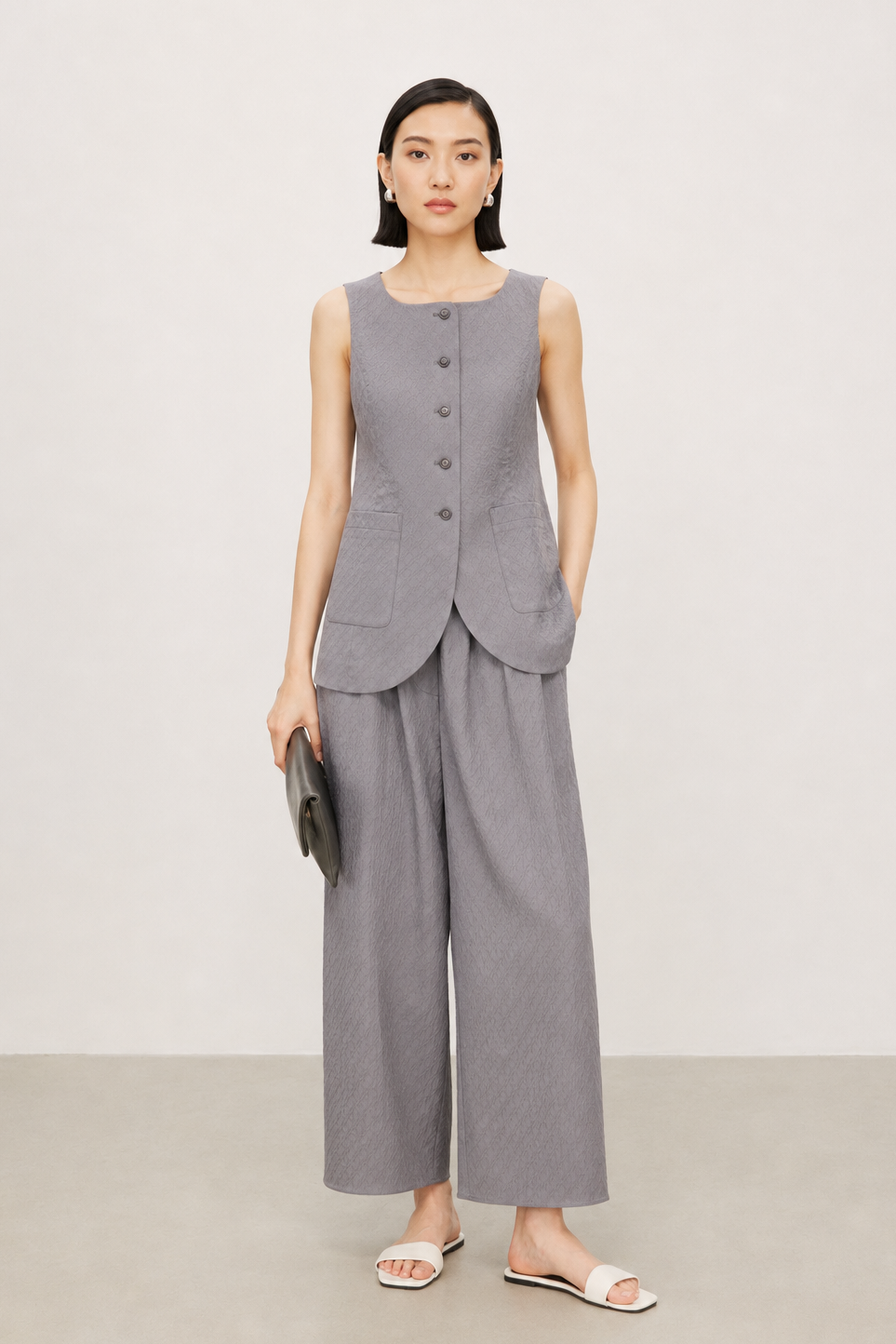 A model stands against a neutral studio backdrop wearing a matching gray sleeveless vest and wide-leg cropped trousers set, holding a small dark clutch and wearing white slide sandals.