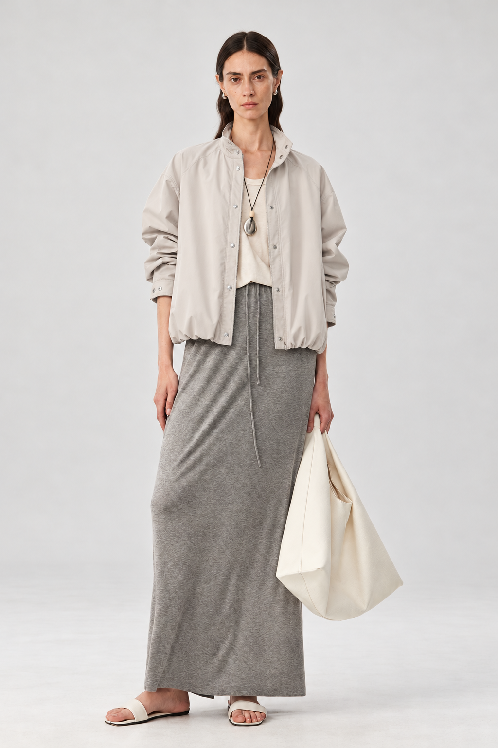 A model stands against a light gray studio background wearing a light beige button-up overshirt over a cream top, a long gray knit maxi skirt, and holding a large cream canvas tote bag.