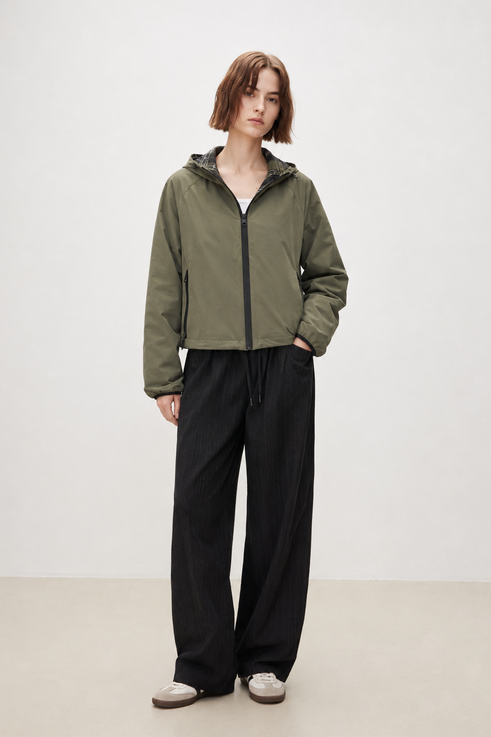 A model stands in a studio wearing an olive green lightweight zip-up hooded jacket, black wide-leg pants, and white sneakers against a plain light gray background.