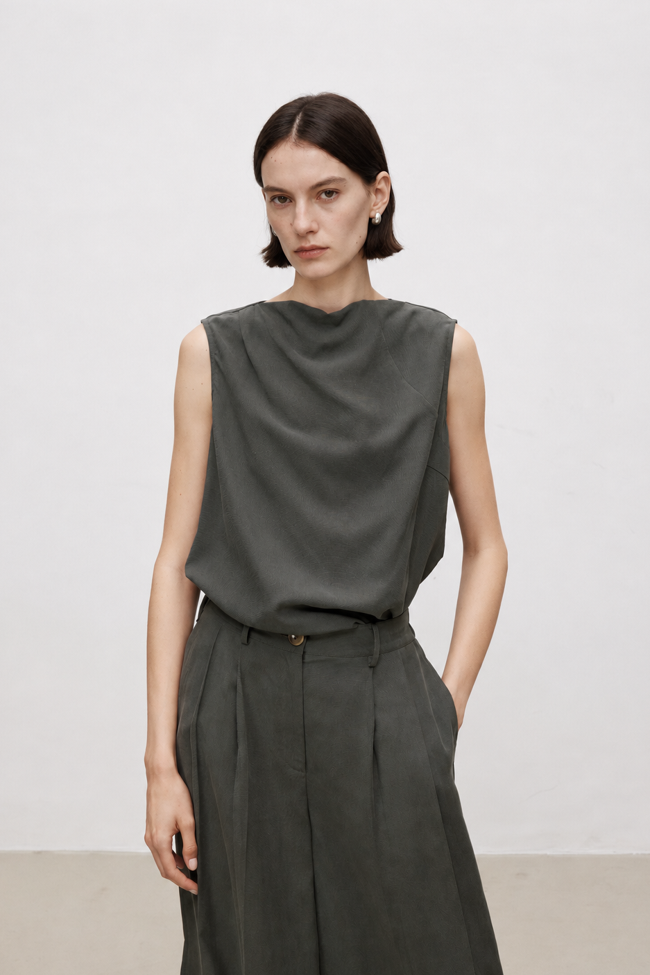A model wearing a dark olive green sleeveless draped top and matching wide-leg trousers, standing against a plain light gray studio wall.