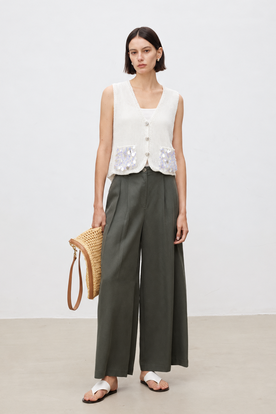 A model stands against a light gray studio wall wearing a sleeveless cream vest with white floral embroidery over dark olive green wide-leg trousers, holding a woven straw bag.