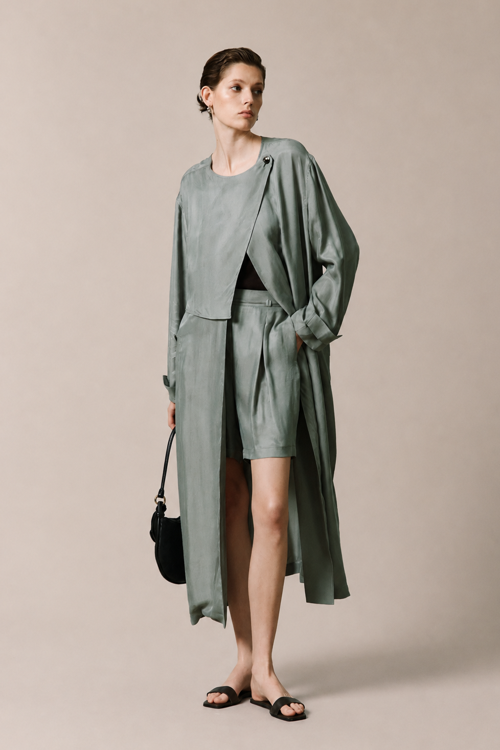 A model stands against a plain beige studio background wearing a matching sage green two-piece outfit consisting of a long draped cardigan and shorts, styled with black sandals and a small black handbag.