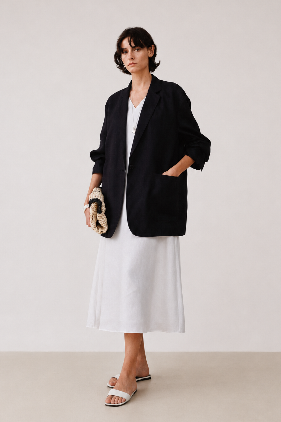 A model stands in a minimalist studio setting wearing an oversized black blazer over a white midi dress, holding a small woven clutch, and wearing white slide sandals.