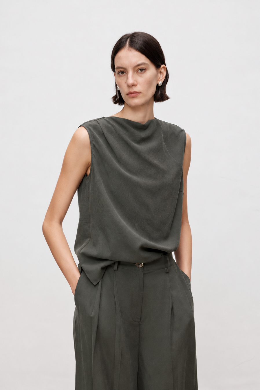 A model wearing a dark olive green sleeveless top with a draped neckline and matching wide-leg trousers, standing against a plain light gray studio background.