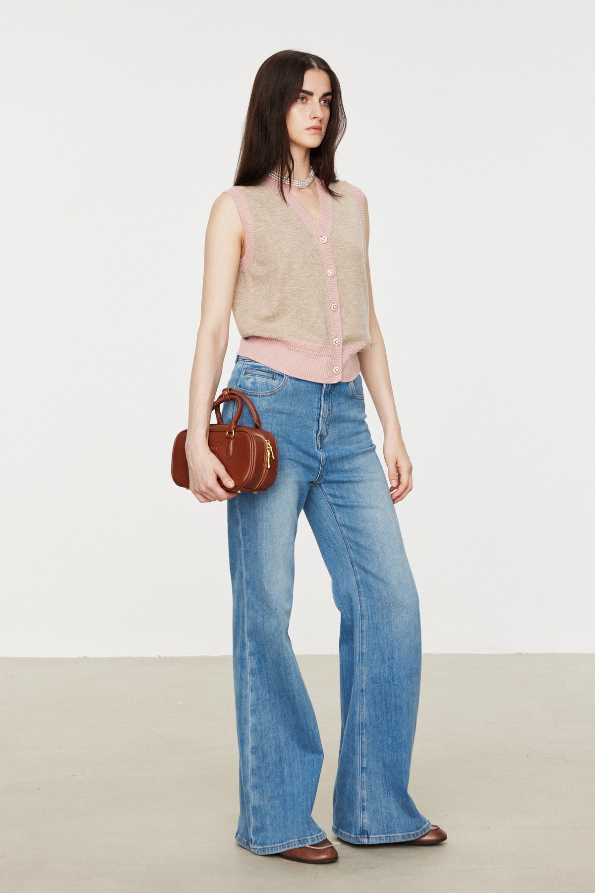 A model stands in a studio wearing a beige sleeveless knit vest over a light pink top, blue wide-leg jeans, and brown shoes, while holding a small brown handbag.