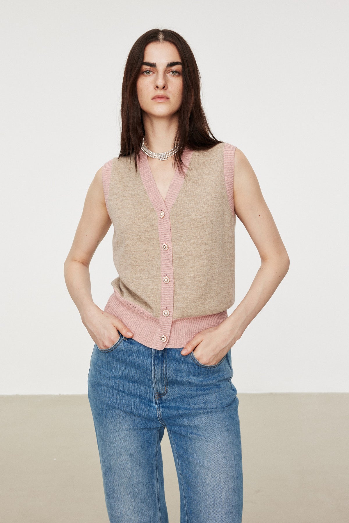 A woman modeling a sleeveless knit vest with a beige body and pink trim, paired with blue jeans, against a plain light gray studio background.