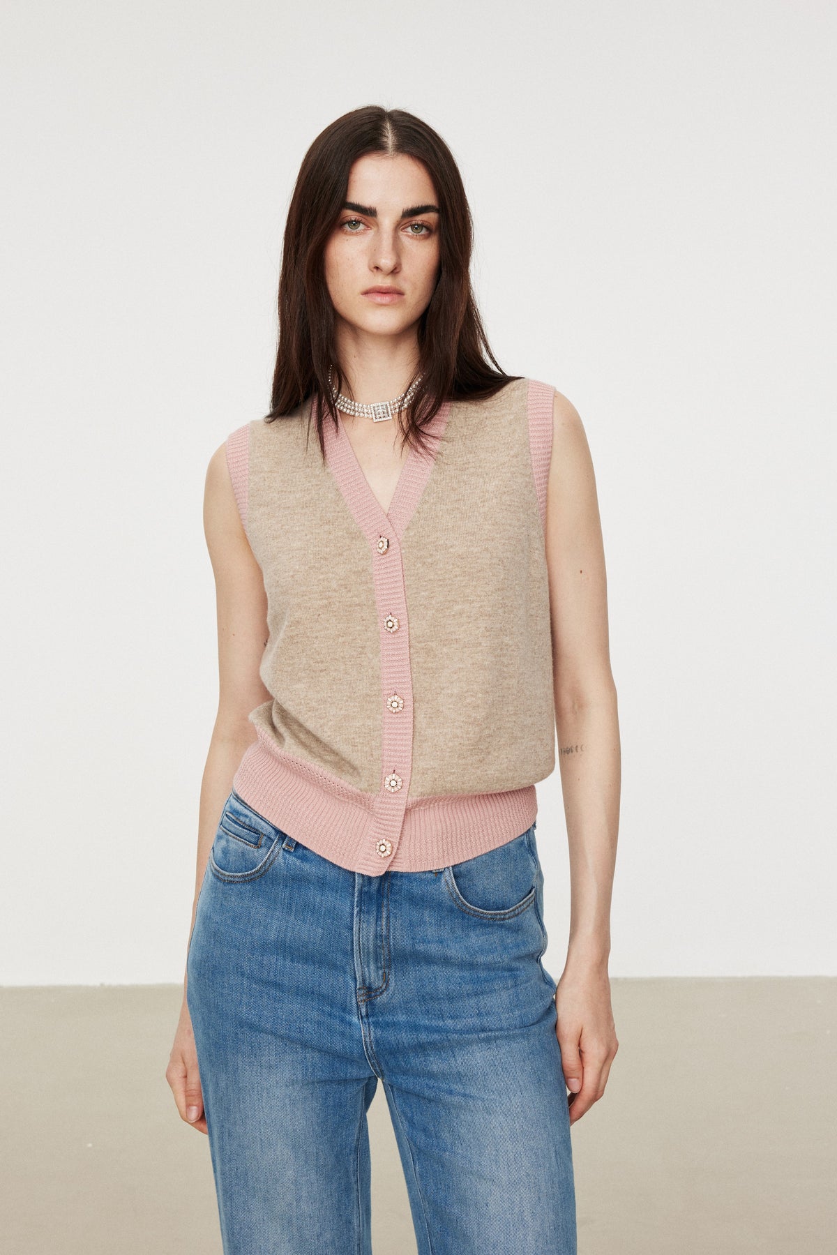A model wearing a beige knit cardigan vest with pink trim and buttons, paired with blue jeans, against a plain light gray studio background.