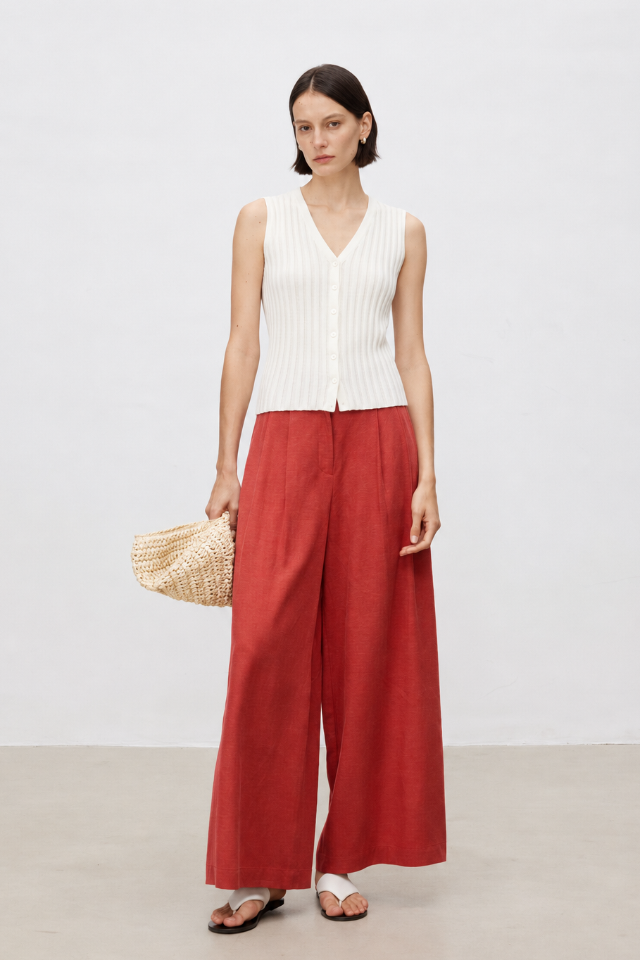 A woman stands in a studio wearing a sleeveless white ribbed knit top and wide-leg rust red trousers, holding a woven straw bag.