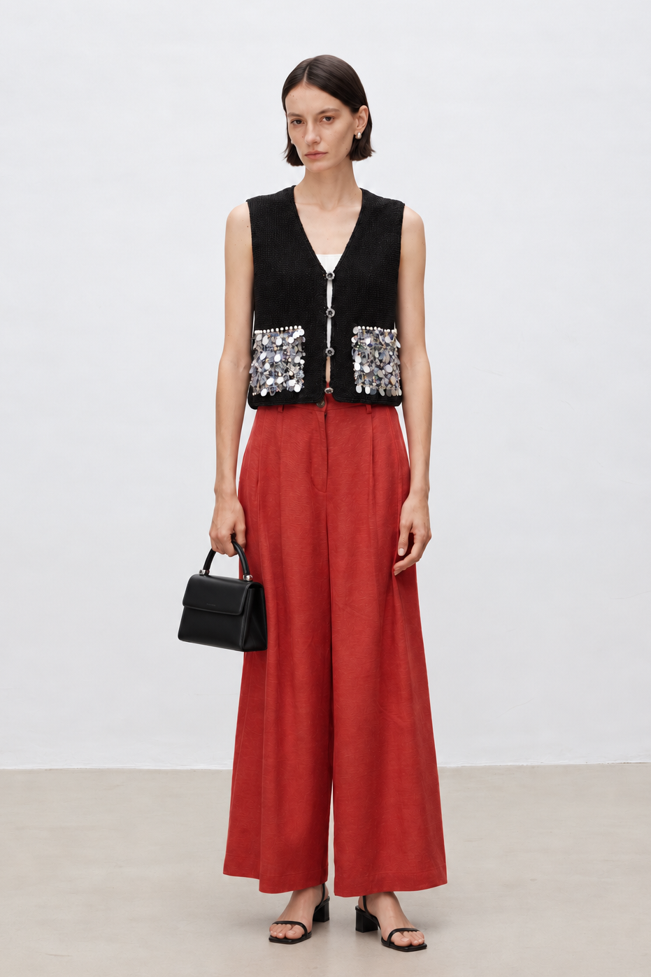 A model stands against a plain white studio background wearing a sleeveless black vest with silver sequin panels at the waist, paired with wide-leg rust-red trousers and black sandals, while holding a small black handbag.