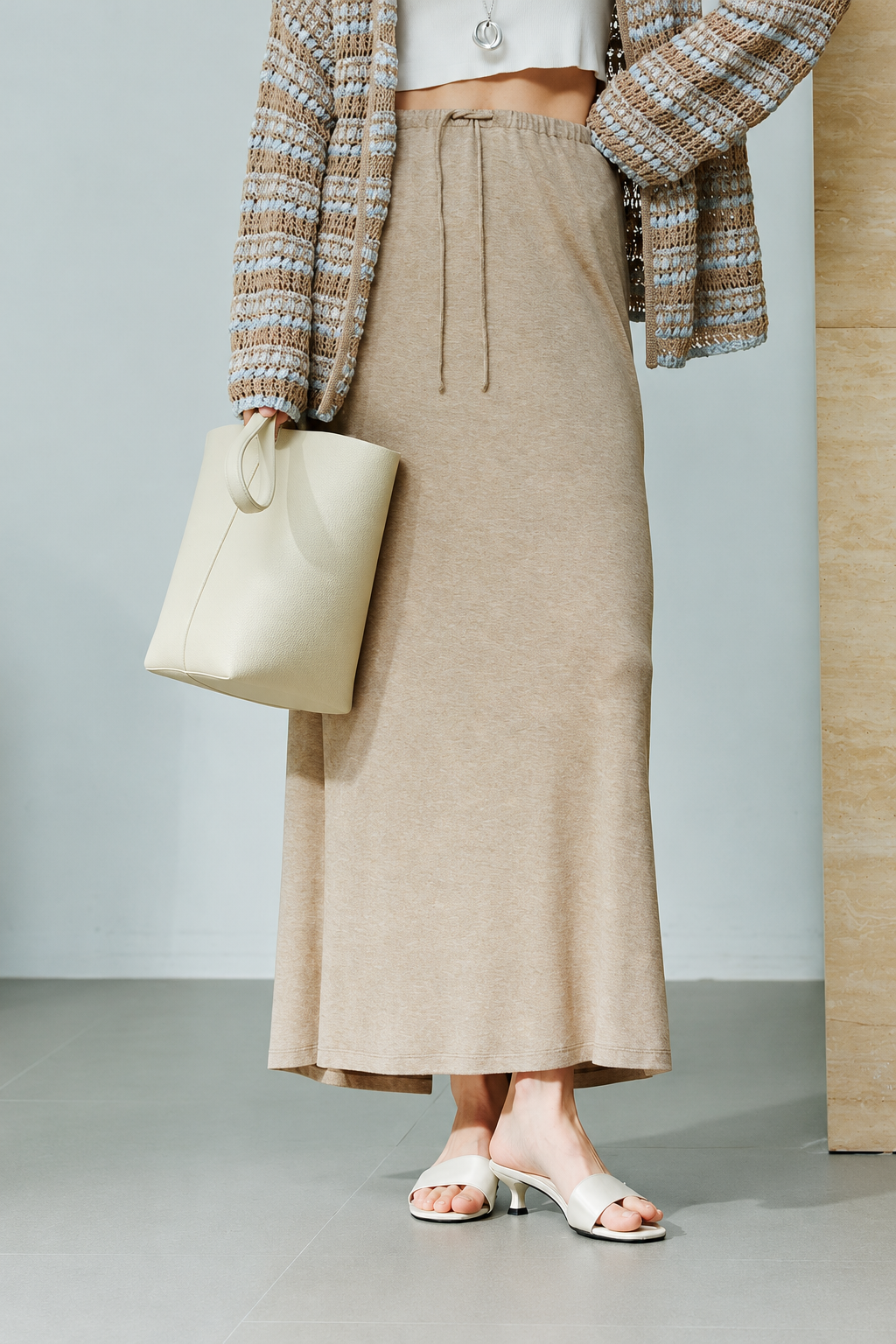 A model is shown from the waist down wearing a beige knit midi skirt with a drawstring waist, styled with a cream-colored knit cardigan, a cream tote bag, and white slide sandals.