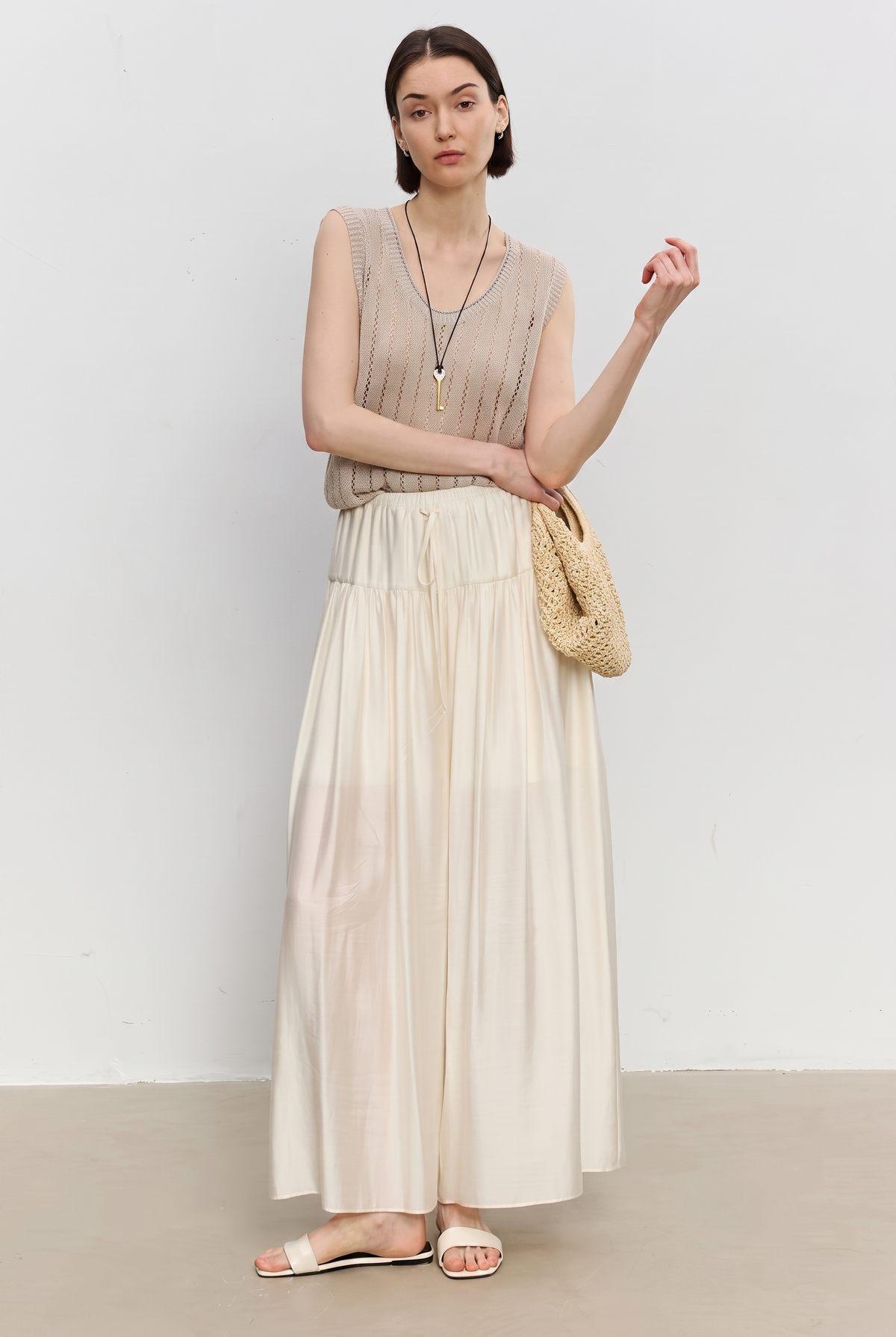 A woman modeling a sleeveless beige top and a long cream pleated skort, holding a woven straw bag, standing against a plain light gray wall.
