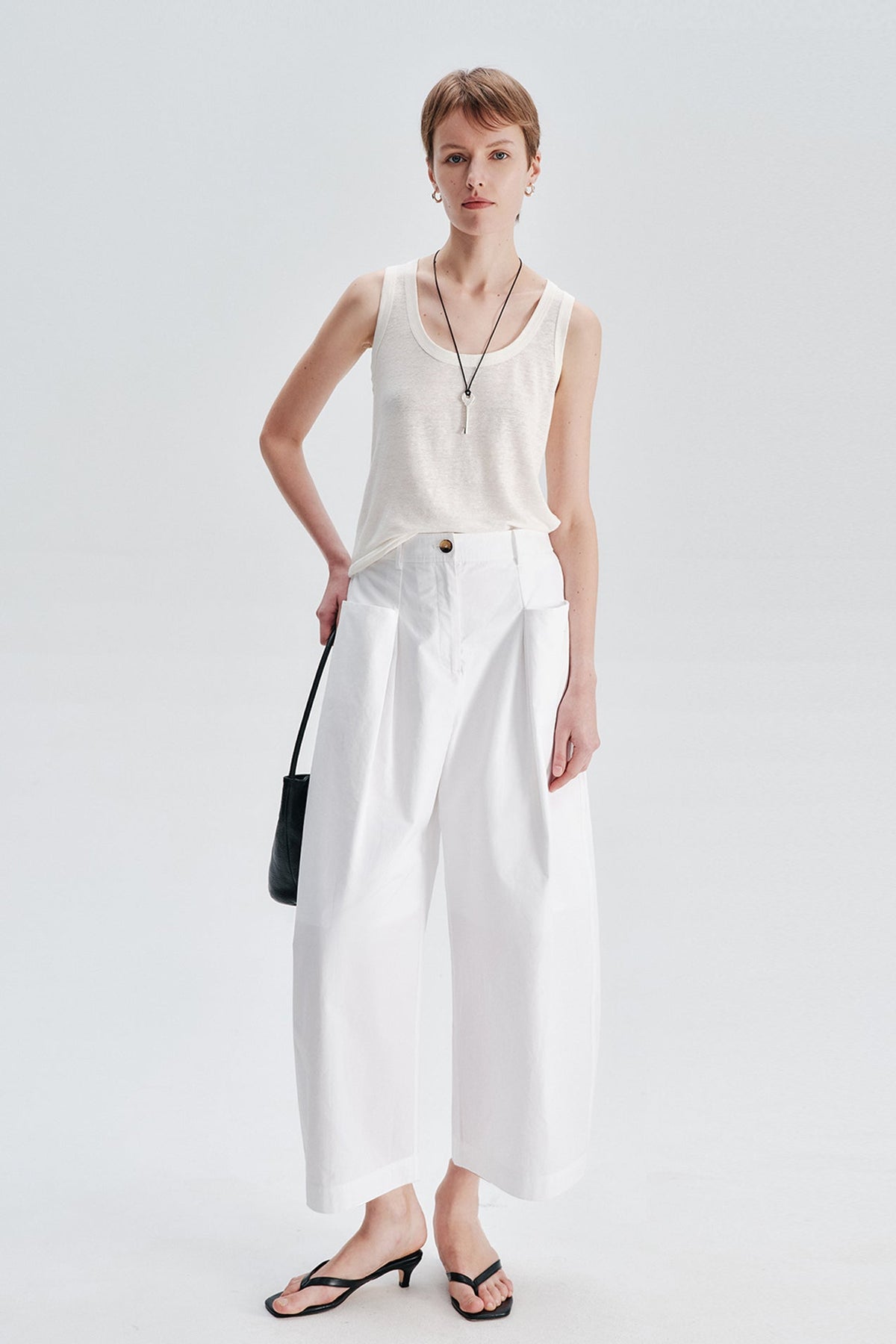 A model stands against a light gray studio background wearing a sleeveless cream top, white wide-leg cropped pants, black strappy sandals, and a small black handbag.