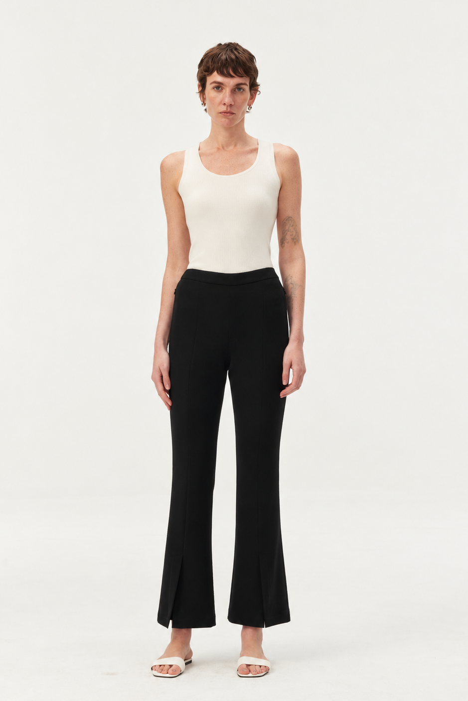 A model stands against a plain white studio background wearing a white ribbed tank top and black wide-leg cropped pants with a slight flare at the hem, paired with white slide sandals.