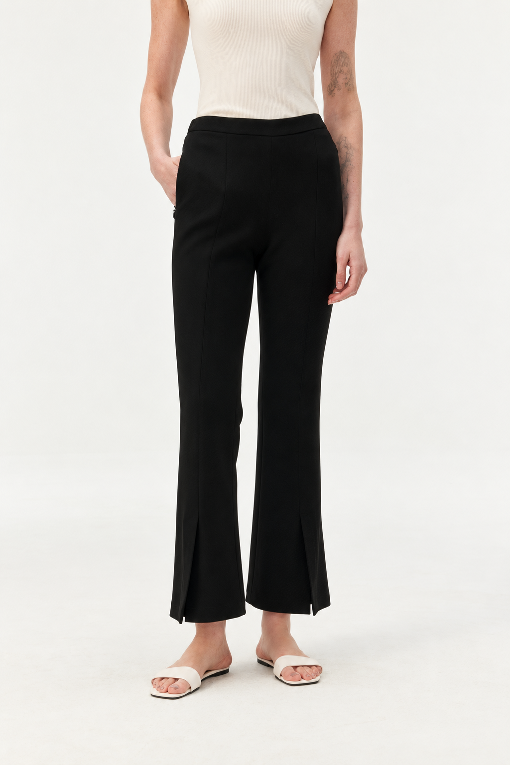 A model wearing black flared cropped pants with front slit details at the hem, paired with a cream sleeveless top and white slide sandals, against a plain white studio background.