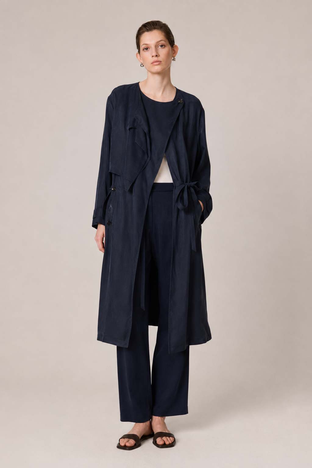 A model stands against a plain beige studio background wearing a dark navy blue longline wrap coat over a matching navy top and wide-leg navy trousers, with black slide sandals.