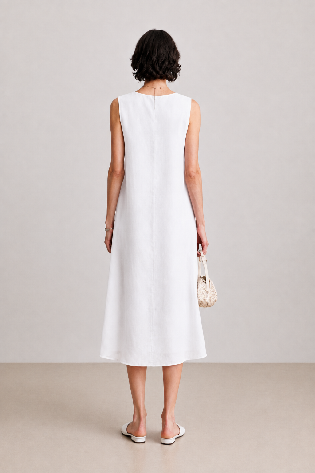 A woman viewed from behind wearing a sleeveless white midi dress, holding a small beige handbag, against a neutral studio backdrop.
