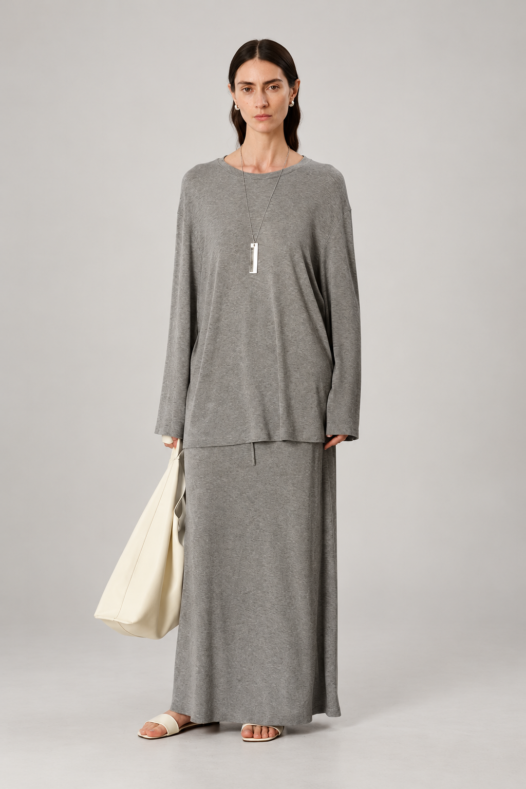 A model stands against a light gray studio background wearing a long-sleeve gray knit top and matching maxi skirt set, holding a cream-colored tote bag.