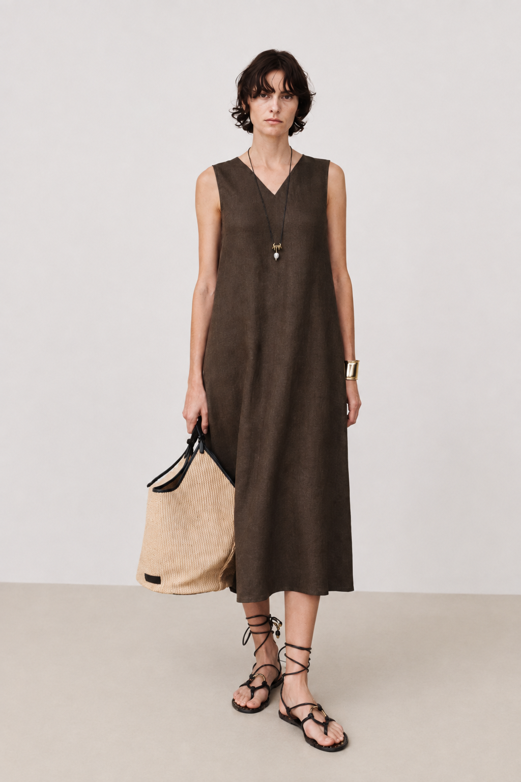 A woman models a sleeveless, V-neck midi dress in a dark brown linen-like fabric, holding a beige tote bag. She is wearing black lace-up flat sandals and a long necklace, standing against a neutral light gray studio background.
