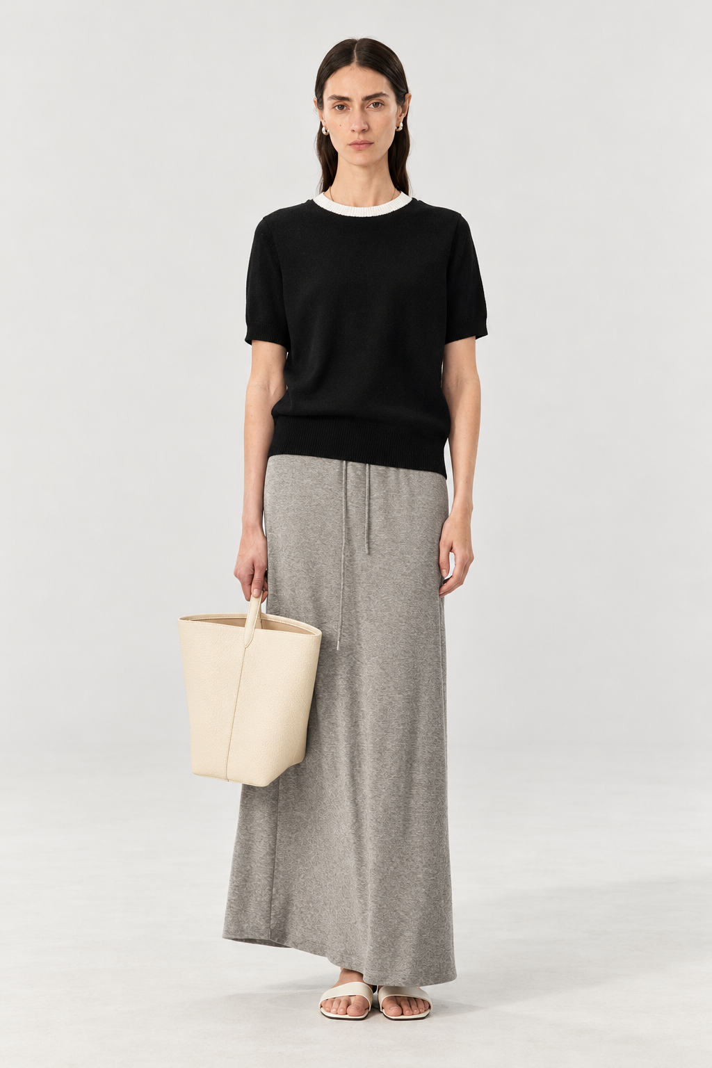 A model stands against a light gray studio background wearing a black short-sleeve knit top with a white crew neck, a long gray drawstring skirt, and holding a cream-colored structured tote bag.