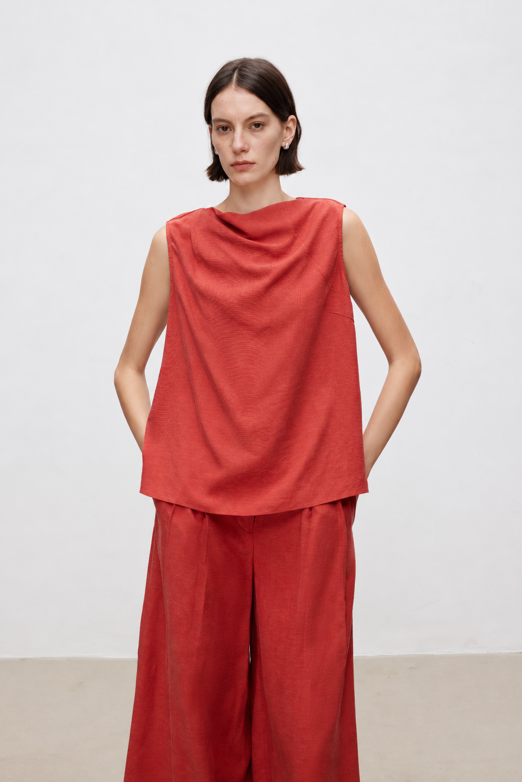 A model wearing a rust-red sleeveless draped top and matching wide-leg trousers, standing against a plain light gray studio background.