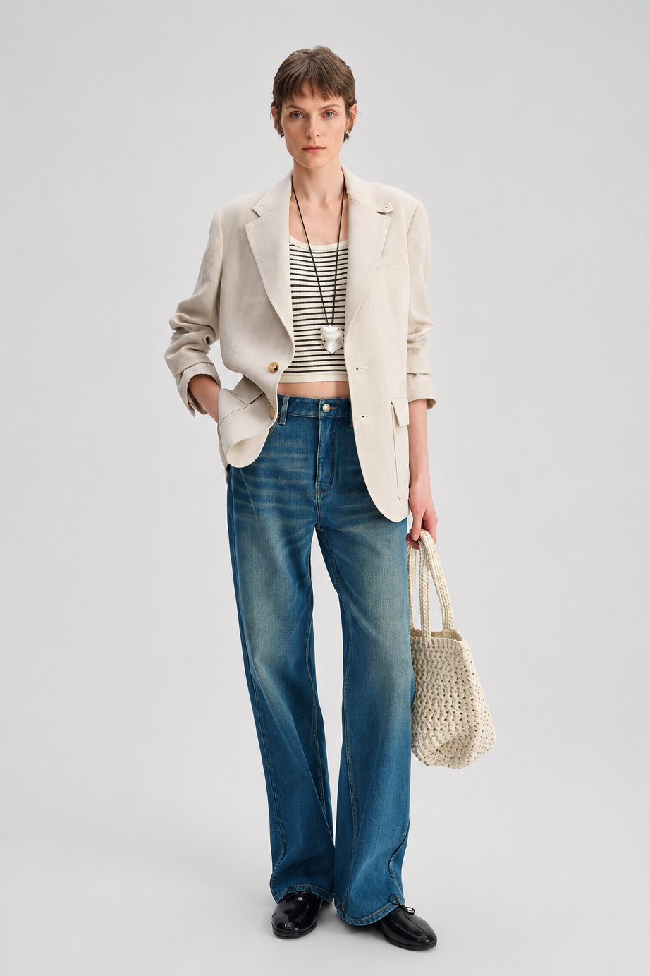 A model stands against a light gray studio background wearing a beige blazer over a black-and-white striped crop top, blue wide-leg jeans, black shoes, and holding a woven straw tote bag.