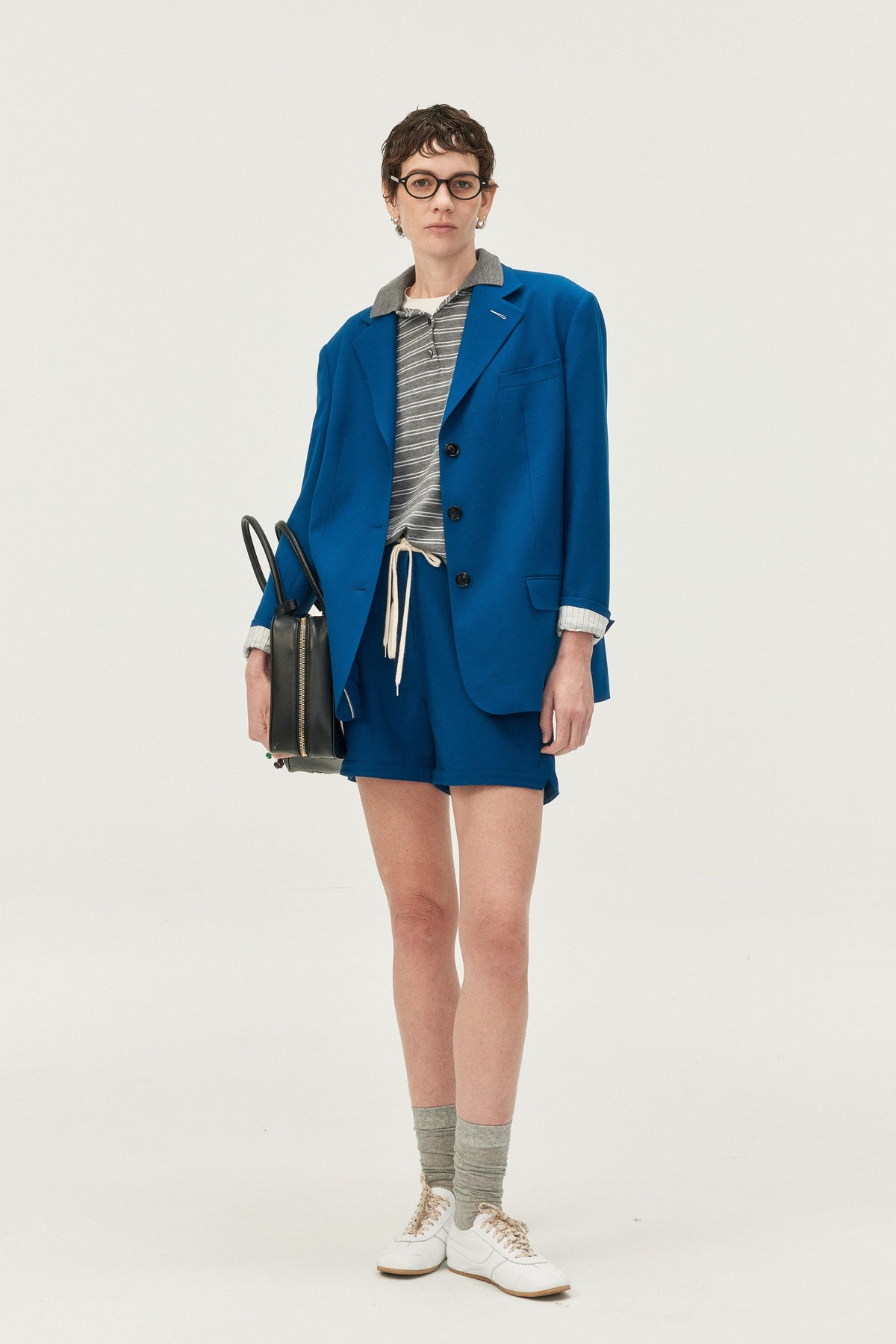 A model stands against a plain light gray studio background wearing a tailored blue blazer and matching shorts over a gray striped top, with white sneakers and a black handbag.