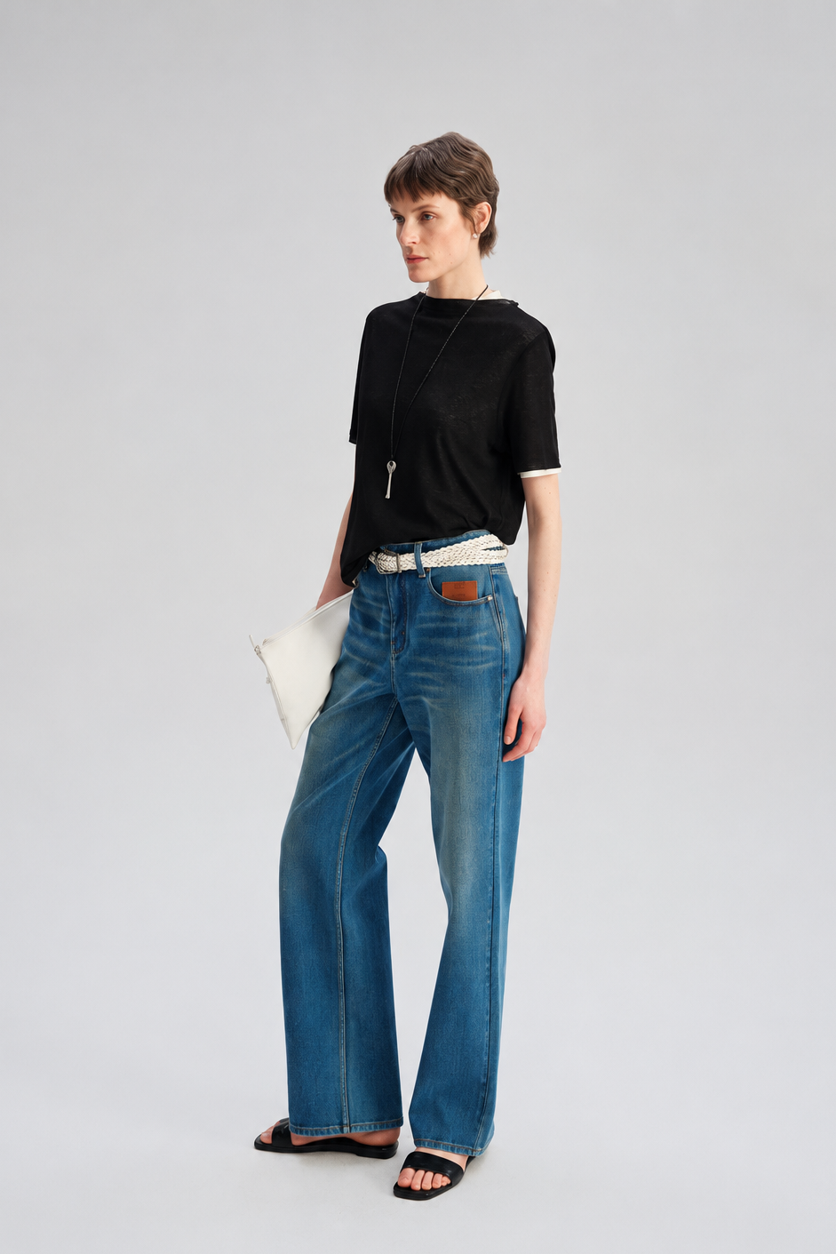 A model stands against a light gray studio background wearing blue wide-leg jeans, a black short-sleeve top, a white belt, black sandals, and holding a white clutch.