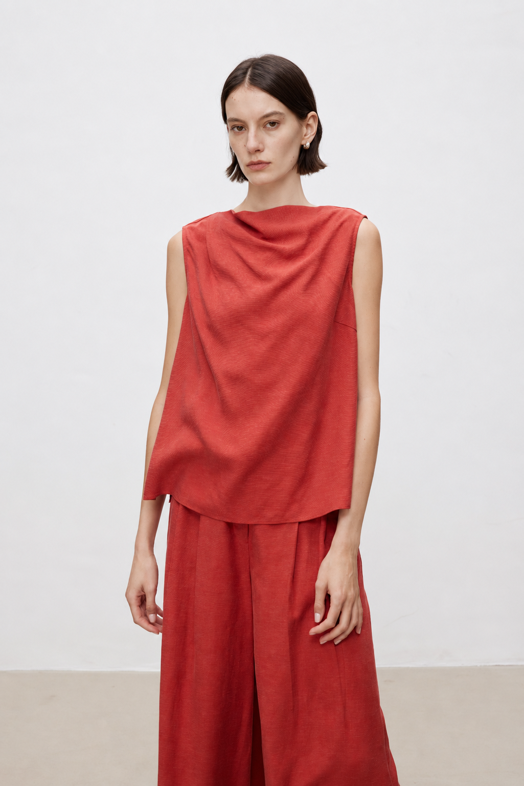 A model wearing a rust-red sleeveless draped top and matching wide-leg trousers, standing against a plain light gray studio background.