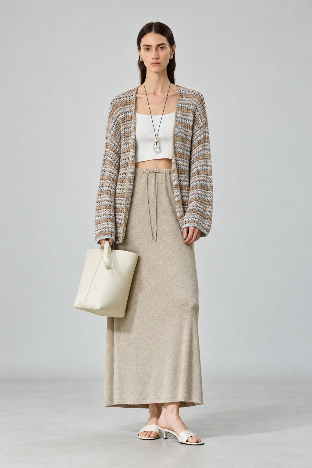 A model stands against a light gray studio background wearing a cream crop top, a long beige knit skirt, a multicolor striped cardigan, and white heeled sandals, while holding a cream tote bag.