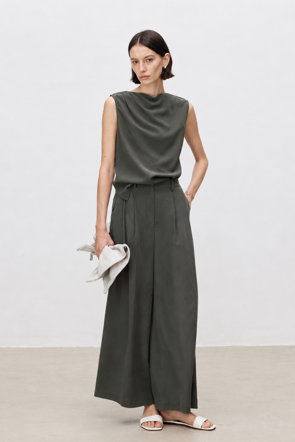 A woman modeling a dark olive green two-piece set consisting of a sleeveless draped top and matching wide-leg trousers, styled with white slide sandals and holding a light-colored clutch against a minimalist studio background.