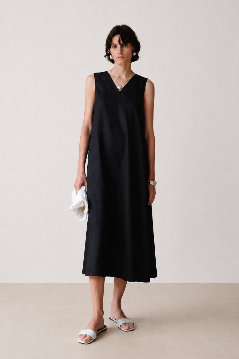 A woman stands in a studio wearing a sleeveless black midi dress with a V-neck, holding a small white textured clutch and wearing white slide sandals. The background is a clean, neutral beige studio wall and floor.