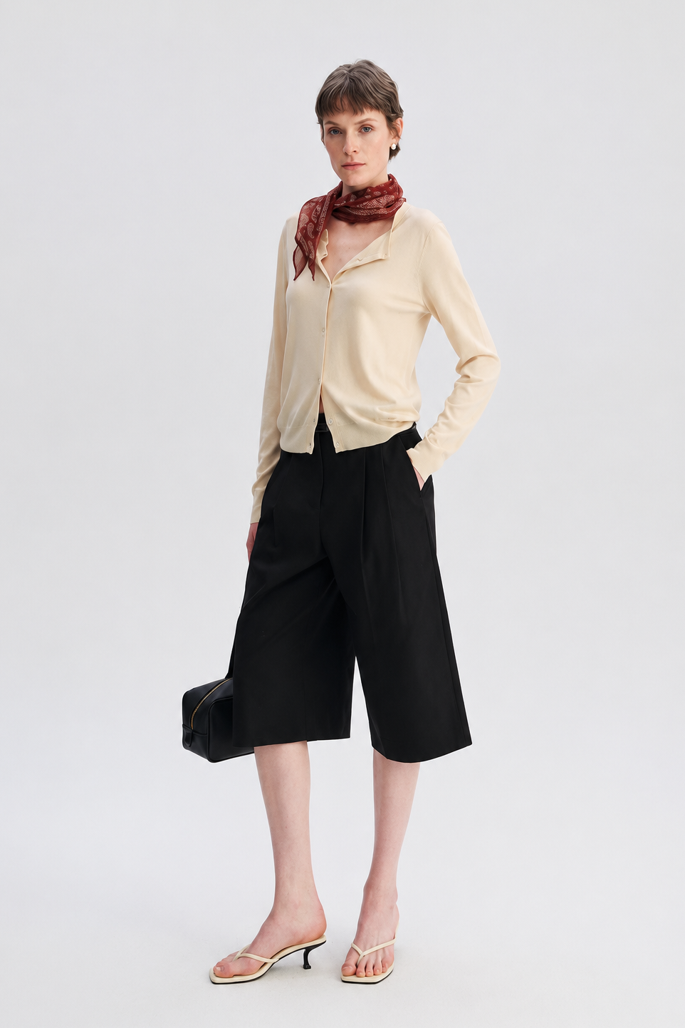 A model stands against a light gray studio background wearing a cream long-sleeve button-up shirt, black wide-leg cropped pants, a red patterned scarf, and clear-strap heeled sandals.