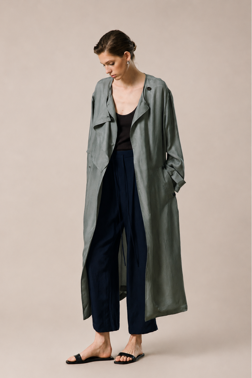 A woman models a long, sage green linen-blend trench coat over a black top and navy blue cropped trousers, standing against a solid beige studio background.